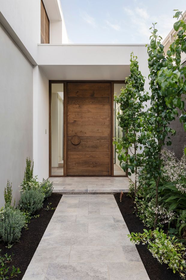 A refined residential entrance courtyard viewed from the garden path. Large pale stone or concrete-look paving leads to a substantial horizontally timber-clad pivot door framed by dark timber or metal glazing. White rendered walls, a flat roofed canopy, and a screened upper-level opening create a clean contemporary facade. Carefully planted garden beds line both sides of the path, adding a lush natural contrast to the minimalist architecture.
