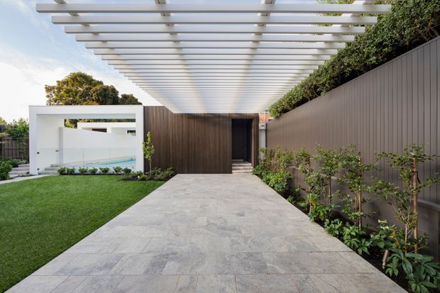 A modern landscaped entry courtyard with a broad pale stone-paved path leading beneath a long white pergola to a dark timber-clad entrance. A manicured lawn, planted garden beds, tall boundary fencing, and a partially visible glass-fronted structure frame the approach.