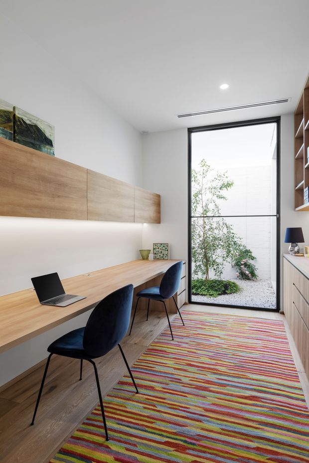 A bright, compact contemporary home office with a long built-in timber desk and overhead timber cabinetry, two dark upholstered chairs, a laptop, and open shelving. A full-height black-framed glazed door opens to a small landscaped courtyard. The room has white walls, light timber flooring and joinery, recessed lighting, and a colourful striped rug.