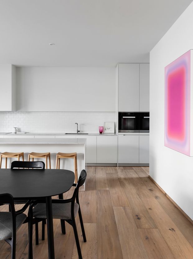 A bright, minimalist open-plan kitchen and dining area. The kitchen features white flat-panel cabinetry, integrated black appliances, a white subway-tile splashback, and a slim island with timber bar stools. A black oval dining table and black chairs occupy the foreground, while wide natural timber flooring and a vivid pink abstract artwork add contrast.