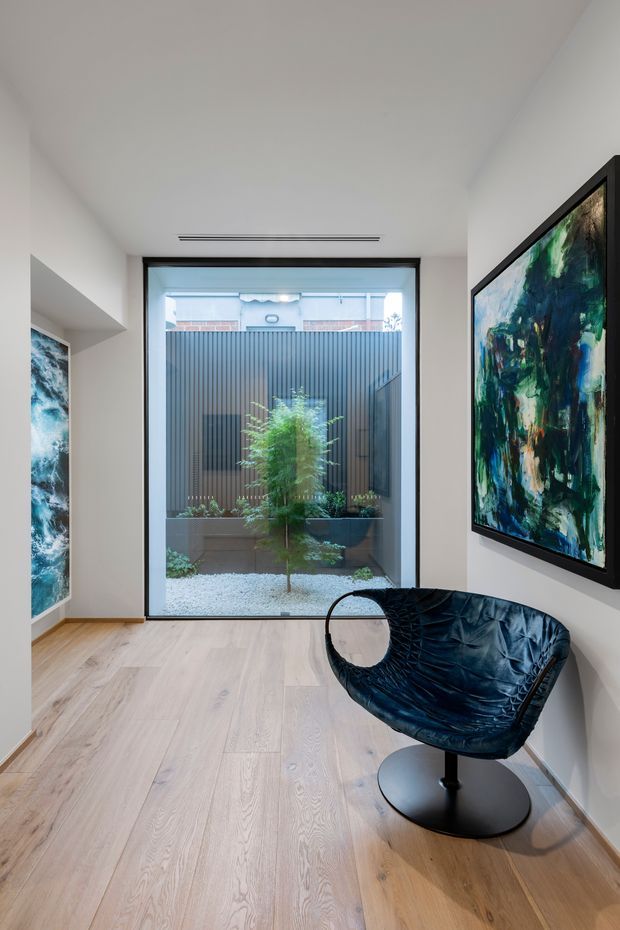 A bright, minimalist interior hallway or gallery-like sitting area with pale timber flooring, white walls, black-framed full-height glazing, and a small planted courtyard visible beyond. Framed abstract artworks hang on both side walls, while a sculptural black leather lounge chair sits in the foreground. A slim linear ceiling vent adds a discreet contemporary detail.