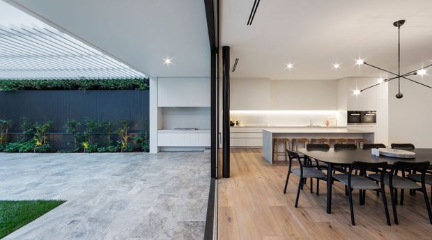 A contemporary open-plan kitchen and dining area opening directly onto a landscaped courtyard through large glazed folding or sliding doors. The interior features a light timber floor, white walls and ceiling, a minimalist white island with integrated cabinetry, pale benchtops, and a black dining table with matching chairs. The courtyard has large-format pale paving, a dark privacy wall, planted garden beds, and a translucent overhead pergola or roof structure. Linear recessed lighting and a sculptural black pendant add modern detailing.