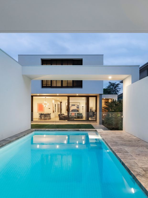 A contemporary two-storey house arranged around a rectangular swimming pool and paved courtyard. The image looks from the pool toward a white rendered facade with a broad cantilevered upper-level frame, large glazed sliding doors, dark horizontal window shading, and warm interior lighting. A dining area and living room are visible through the glazing, with landscaped planting along one side. The scene is photographed at dusk and has a clean, minimalist architectural character.