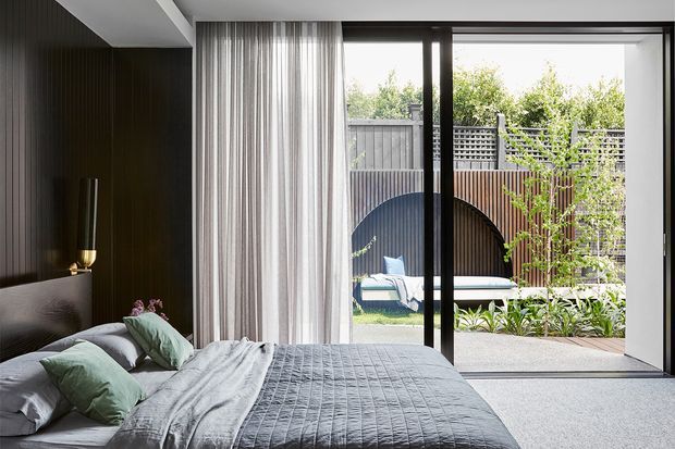 A contemporary bedroom with a low upholstered bed dressed in grey bedding and muted sage cushions. The room features a dark vertically ribbed feature wall, sheer full-height curtains, and large black-framed sliding glass doors opening to a landscaped private courtyard with timber screening and an outdoor daybed. The palette is restrained and cool-neutral, with strong indoor-outdoor connection and abundant natural light.
