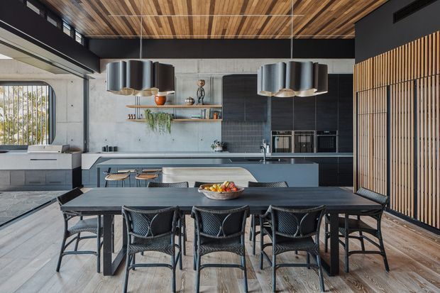 A contemporary open-plan kitchen and dining area with a long dark dining table and woven dining chairs in the foreground. The kitchen features a broad island with integrated seating, dark blue-grey cabinetry, a concrete-look wall, open timber shelving, integrated appliances, and multiple sculptural pendant lights. A timber-lined ceiling and vertical timber screen add warmth and texture, while large glazing and pale flooring keep the space bright and airy.