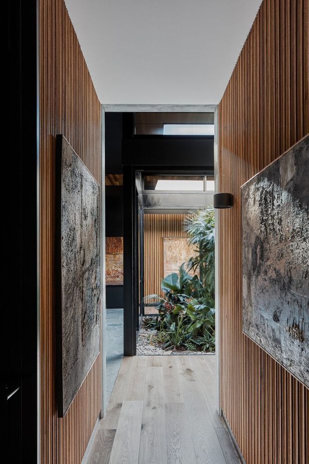 A narrow, refined interior passage lined with vertical timber battens and large textured dark stone feature panels. Pale timber flooring leads toward a glazed opening and planted courtyard, with black-framed glazing, dark ceilings, and a skylight or clerestory bringing in natural light. The composition emphasizes material contrast, strong vertical rhythm, and an indoor-outdoor connection.