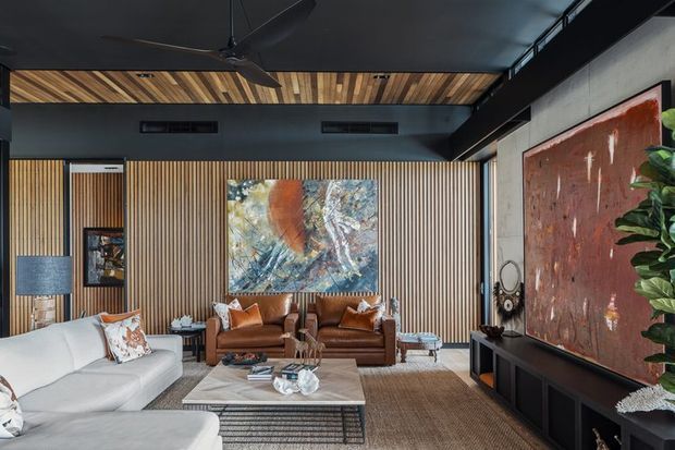 A sophisticated contemporary living room with a large white sectional sofa, cognac leather seating, a low rectangular coffee table, and a dark built-in media console. Vertical timber slat wall panelling, a timber-lined ceiling section, dark painted surfaces, exposed concrete, large abstract artwork, and a ceiling fan create a warm yet dramatic interior.