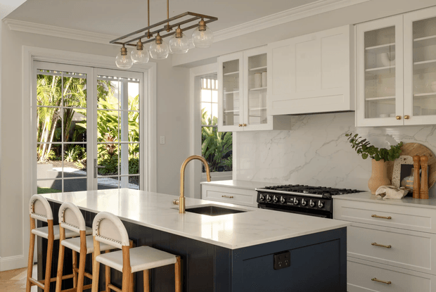 Bright, refined kitchen with a large white stone island, dark navy island cabinetry, white shaker-style perimeter cabinetry, brass hardware and faucet, and a marble-look splashback. The room features a glazed external door opening to greenery, a freestanding black range cooker, rattan bar stools, and a linear pendant light with clear glass globes.