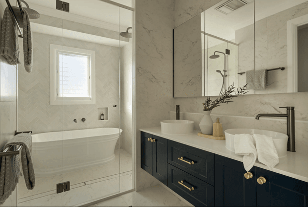A refined contemporary bathroom featuring a freestanding white bathtub enclosed by clear glass, a large mirrored wall, and a double vanity with dark blue cabinetry, brass hardware, a pale stone benchtop, and vessel basins. Light marble-look wall surfaces, herringbone tile detailing, black tapware, and soft neutral textiles create a bright, elegant interior.