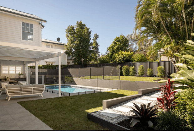 A landscaped residential backyard with a rectangular swimming pool, paved patio, covered outdoor seating area, manicured lawn, raised garden beds, and tropical planting. The two-storey home has pale horizontal weatherboard cladding, a light metal roof, large windows with shutters, and a contemporary covered entertaining structure. Black fencing and dark pool edging contrast with the light paving and greenery.