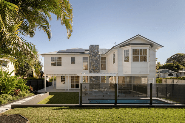 Exterior view of a light-coloured, multi-level suburban residence with white weatherboard cladding, a prominent stone chimney, pitched gable roofs, solar panels, and large glazed windows. A fenced swimming pool and landscaped lawn occupy the foreground, with tropical planting at the side.