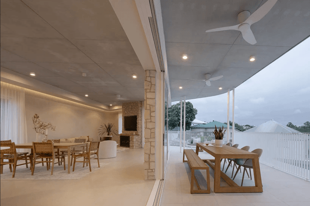 A contemporary indoor-outdoor dining area with large fully open glass doors connecting a warm, softly lit interior dining and living space to a covered outdoor terrace. The interior features a timber dining table and chairs, pale neutral finishes, a stone-clad fireplace wall, recessed lighting, and sheer curtains. The terrace has a timber dining setting, ceiling fans, slender posts, and white balustrading overlooking neighbouring rooftops and trees.