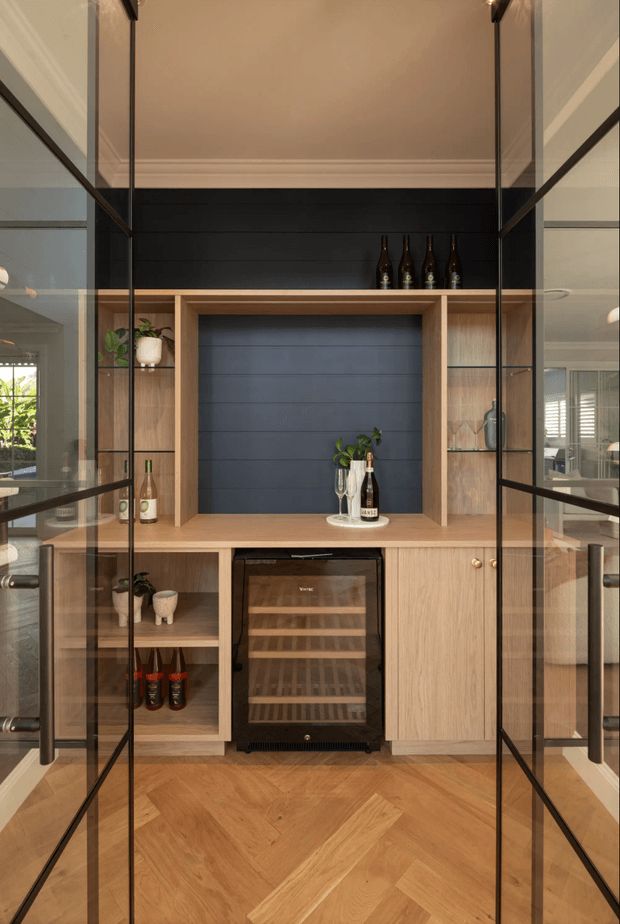 A built-in home bar and wine storage joinery viewed through framed glass doors. The cabinetry is warm light timber with open shelving, a dark charcoal painted feature backing, integrated wine refrigerator, bottle storage, decorative bottles, and small plants. The floor is laid in a warm timber herringbone pattern, while the surrounding glass and black metal framing create a refined, contemporary display-room feel.
