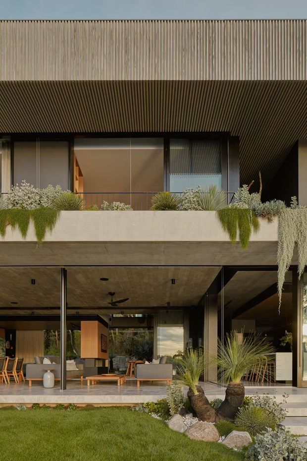 A modern multi-level residential facade with deep roof overhangs, extensive glazing, vertical timber-look screening and a covered outdoor living area. The open ground floor contains lounge furniture, while lush drought-tolerant planting, raised planters, rocks and lawn create a landscaped setting. Warm neutral materials and strong horizontal and vertical lines define the composition.