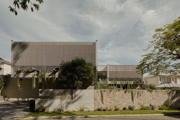 Street-facing view of a contemporary multi-level residential development with vertically ribbed metal-clad upper volumes, pale rendered lower walls, landscaped frontage, and a rough stone retaining wall. Mature planting and neighbouring homes frame the composition.