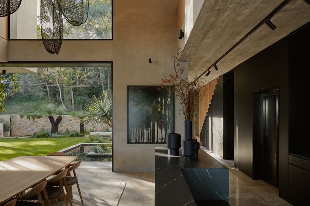 A refined open-plan dining and living space with a long timber dining table beside expansive glazing overlooking a landscaped garden. The interior features warm textured plaster walls, a large dark stone island or console, black built-in cabinetry, a concrete-look ceiling with track lighting, and a sculptural open-riser staircase. Floor-to-ceiling windows and a glazed door create a strong connection to the garden, while pendant lights and dark decorative vessels add a contemporary, earthy character.