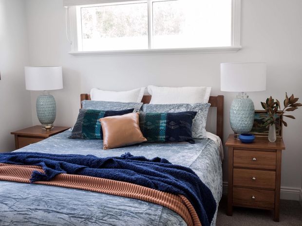 A bright, neatly styled bedroom with a timber bed, layered white and blue bedding, navy throw, and copper-toned accent cushion. Matching timber bedside tables flank the bed, each with a textured blue ceramic lamp and white shade. A large white-framed window provides natural light, while a small plant adds warmth.