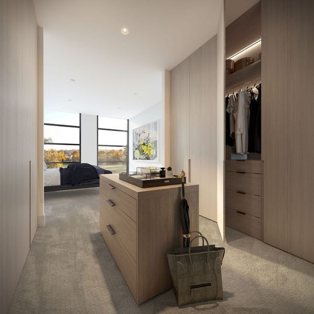 A bright, contemporary bedroom dressing area with a central wood-finished island dresser, floor-to-ceiling built-in wardrobes and an open hanging section. The bedroom is visible beyond the dressing zone, with a dark upholstered bed, large black-framed windows, artwork, and a soft grey carpet. Integrated wardrobe lighting and concealed ceiling lighting create a clean, refined interior.