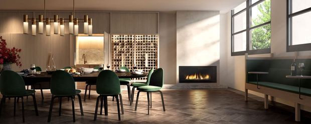 A sophisticated open-plan dining and living area with a long dark dining table surrounded by deep green upholstered chairs. A linear fireplace is set into a pale textured wall, while large black-framed windows bring in natural light and greenery. A sculptural multi-pendant chandelier hangs above the dining table, and a built-in dark green sideboard runs beneath the windows. The floor is laid in a herringbone timber pattern, with a decorative geometric screen and softly illuminated feature wall adding architectural interest.