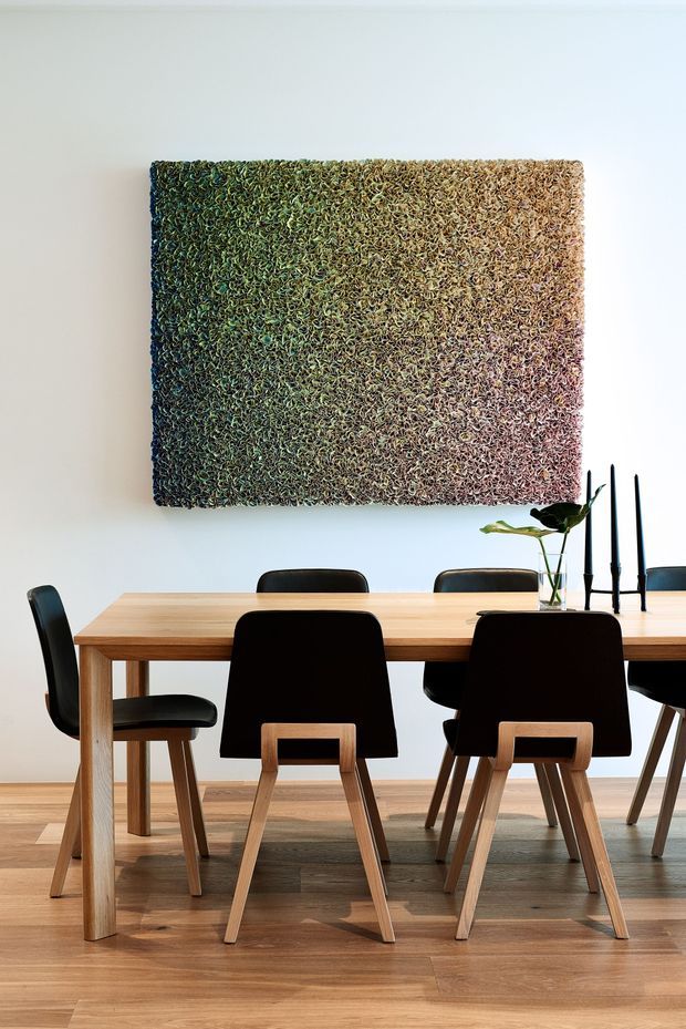 A bright, minimalist dining room featuring a long light-wood dining table with black dining chairs, set against white walls and warm timber flooring. A large textured abstract wall artwork in green, beige, and muted pink tones hangs above the table, with simple black candleholders and a small vase providing understated decor.