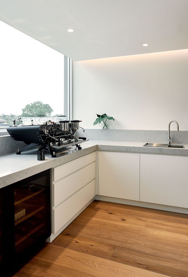 A bright, minimalist kitchen with an L-shaped arrangement of pale cabinetry and a light grey stone benchtop. A large espresso machine sits on the counter beside a dark built-in wine storage unit, while a stainless steel sink and curved faucet occupy the opposite run. Warm timber flooring contrasts with the white walls and cabinetry. Full-height glazing brings in natural light and views of greenery outside, with recessed ceiling lights and concealed warm cove lighting.