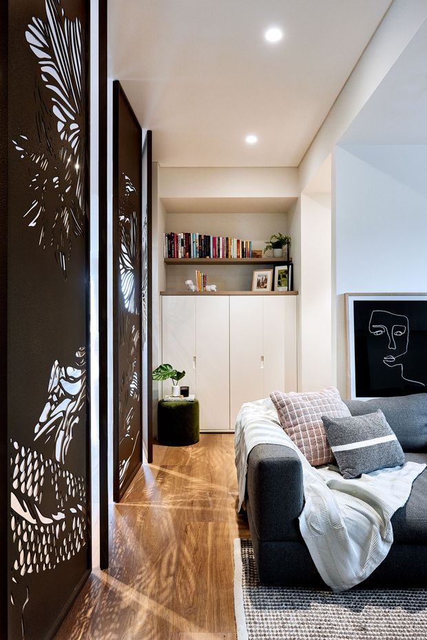 A contemporary living room with a dark sectional sofa, layered neutral throws and cushions, timber flooring, built-in white cabinetry and open shelving, and a large black-and-white line-art print. Decorative laser-cut dark metal screens with botanical patterns create a striking partition along the left side, casting patterned shadows across the floor. Recessed ceiling lights and a warm neutral palette give the room a refined, inviting atmosphere.