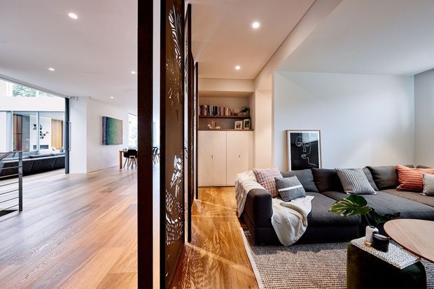 Open-plan contemporary interior showing a living room in the foreground with a large sectional sofa, low coffee table, built-in cabinetry and warm timber flooring. Decorative timber screens partially divide the living area from a bright adjacent space, with further seating and glazing visible beyond.