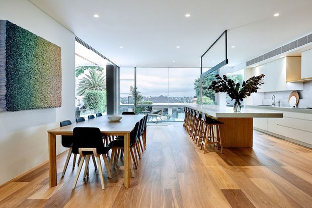 A bright, open-plan contemporary kitchen and dining area with warm timber flooring, a timber dining table, black dining chairs, and a large white kitchen with a timber island and bar seating. Full-height glazing opens to a leafy view and distant water, while a green textured wall artwork adds a natural accent. Recessed ceiling lights and a linear air-conditioning vent contribute to the clean modern fit-out.