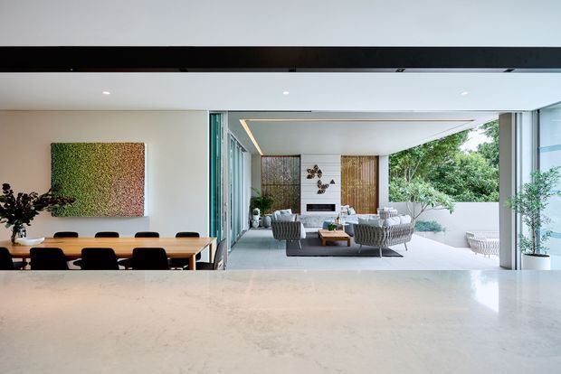 Open-plan contemporary living and dining area viewed across a light stone island or benchtop. The dining zone has a long timber table with black chairs, while the adjoining lounge features grey seating, a coffee table and large sliding glass openings to a planted outdoor terrace. The space is bright and airy with extensive glazing, white walls and integrated indoor-outdoor flow.