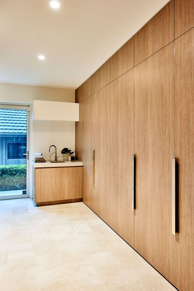 A contemporary utility or laundry space with full-height light timber cabinetry, long vertical handles, a compact sink and tap, pale stone-look flooring, and a small window/door opening to the exterior. The room is clean, warm, and highly integrated, with concealed storage dominating one wall.