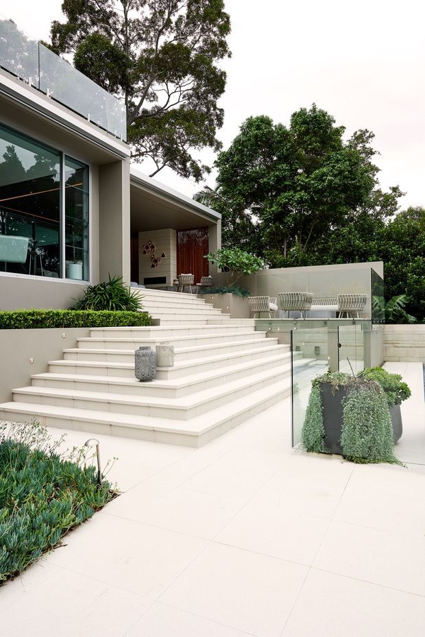 A contemporary multi-level outdoor terrace and garden surrounding a modern house. Broad pale tiled steps lead to a covered patio with outdoor seating, while glass balustrades, rendered walls, dense planting, and mature trees frame the space. Large glazed openings connect the terrace to the interior, and the overall setting feels refined, bright, and landscaped.
