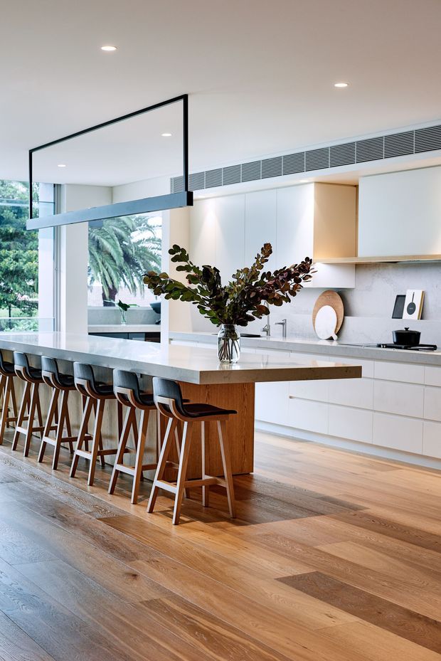 A bright, contemporary open-plan kitchen with a large white island and warm timber base, seating for several people on black upholstered bar stools. White handleless cabinetry, a pale stone benchtop and splashback, integrated linear air-conditioning grille, timber flooring, recessed downlights, and expansive glazing create a clean, refined interior. A sculptural pendant light and vase of foliage add visual interest.