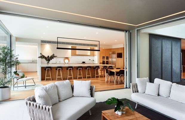 Bright open-plan contemporary home interior viewed from a light-filled living room, with large retractable glazed doors opening to a kitchen and dining area. The space features pale sofas, timber flooring, a white kitchen with island seating, warm timber cabinetry, recessed lighting, and a dramatic dark feature wall near the dining area.