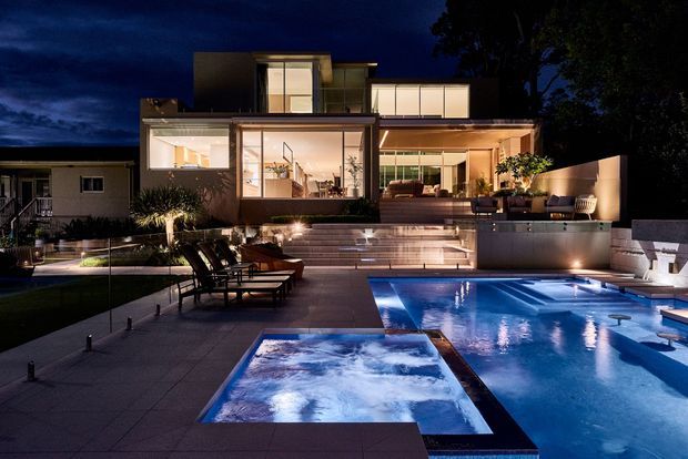 Night-time exterior view of a contemporary multi-level residence with extensive floor-to-ceiling glazing, illuminated interior spaces, layered terraces, and a large backyard pool with an integrated spa or hot tub in the foreground. The landscaped outdoor area includes paved decking, lounge chairs, planting, and architectural lighting.