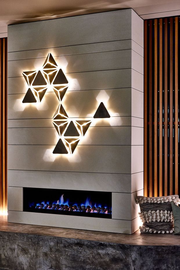 A contemporary interior feature wall centred on a long horizontal electric fireplace with blue flames. The fireplace surround is clad in large-format horizontally jointed grey panels, with sculptural geometric illuminated wall lights arranged above it. A vertical timber slat screen occupies the adjacent wall, while a textured stone hearth or plinth and patterned soft furnishings add contrast.