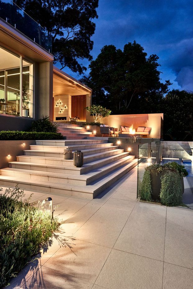 Twilight view of a contemporary residential outdoor entertaining area with broad tiled steps leading to a raised terrace. The terrace includes a fire feature, outdoor lounge seating, integrated lighting, glass balustrades, landscaped planter beds, and a glimpse of a swimming pool. Large glazed openings connect the terrace to the modern interior, while mature trees form the background.