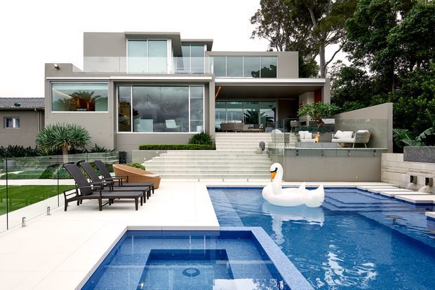 Contemporary multi-level residence with broad glazed openings, flat roof forms, and a stepped terrace overlooking a swimming pool. The pool area includes a shallow square spa or sun shelf, glass balustrading, pale paving, outdoor lounge seating, and a large inflatable swan. Mature trees surround the property.
