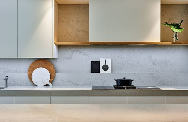 A clean, contemporary kitchen with pale flat-panel cabinetry, a light stone-look splashback and benchtops, integrated cooktop, and warm timber open shelving. Minimal countertop styling includes cutting boards, cookware, and a small plant.
