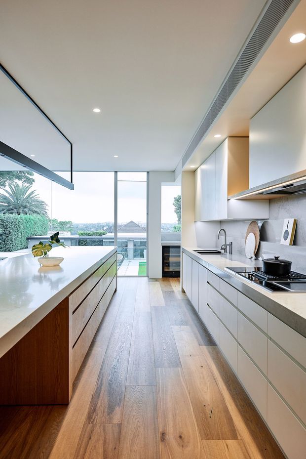 A bright, contemporary galley-style kitchen with a large central island, white stone benchtops, pale built-in cabinetry, integrated appliances, and warm timber flooring. Full-height glazing opens the space to a leafy suburban outlook, while a linear ceiling air-conditioning grille and recessed downlights reinforce the streamlined design.