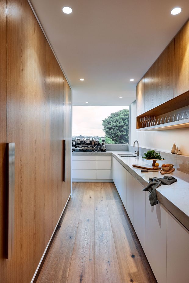 A narrow, contemporary galley kitchen with full-height warm timber cabinetry on the left and sleek white handleless cabinetry on the right. The kitchen features a pale stone benchtop, integrated sink and tap, open timber shelving displaying glassware, a coffee machine, recessed ceiling lights, and wide timber flooring. A large rear window overlooks a leafy suburban setting.