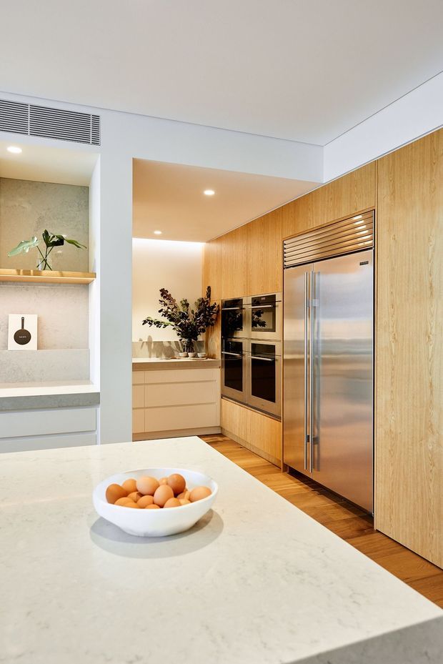 A bright contemporary kitchen with a large pale stone island in the foreground, warm timber full-height cabinetry, integrated stainless-steel double ovens and refrigerator, and a compact rear benchtop. Recessed ceiling lighting, clean white walls, open shelving, and subtle decorative planting create a refined, highly integrated fit-out.