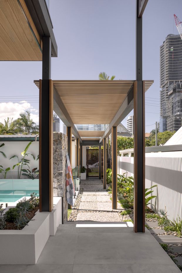 A bright contemporary outdoor passageway beside a residential pool, framed by dark slender posts and a timber slatted pergola. The walkway combines smooth concrete paving, pale gravel, rock-clad planter walls, lush tropical planting, and a turquoise pool. A perforated privacy screen closes the view at the far end, while high-rise construction is visible beyond the property, creating an urban setting.