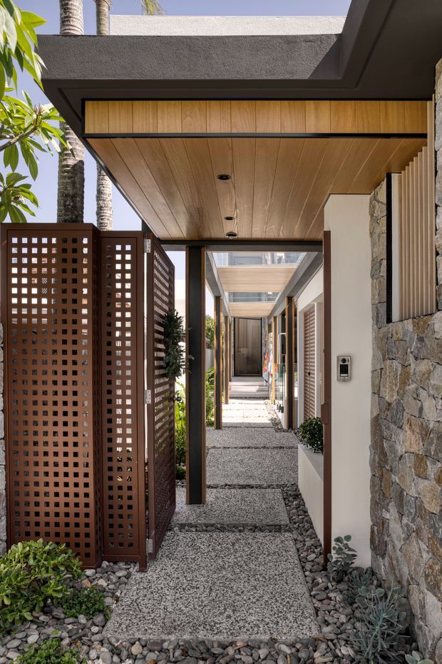 A contemporary covered entry walkway with a textured pebble-finished path, dark river-stone edging, timber-lined soffits, dark structural posts, a perforated metal privacy screen, and a natural rough-stone wall. Tropical planting and strong daylight frame the layered corridor leading toward the entrance.