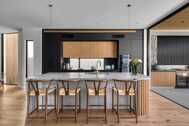 A spacious contemporary kitchen with a large grey stone island and four timber bar stools, dark charcoal cabinetry, and a central timber overhead cabinet. The kitchen features warm timber flooring, vertical timber slat detailing, integrated appliances, pendant lighting, and a secondary kitchen or scullery area visible through the wide opening on the right. The palette is balanced between dark cabinetry, light grey stone, and warm timber.