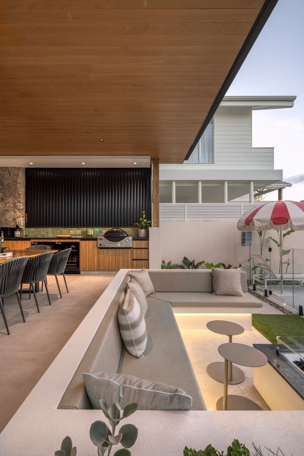 A refined open-plan outdoor entertaining and living area beneath a large timber-lined ceiling. The space includes a built-in outdoor kitchen with timber cabinetry, stone countertop, barbecue, and dark ribbed feature wall, alongside a long integrated upholstered bench, dining table and chairs, and a sunken lounge with layered coffee tables. A glazed opening connects to a bright contemporary coastal-style building, while landscaped planting and a striped outdoor umbrella add relaxed resort character.