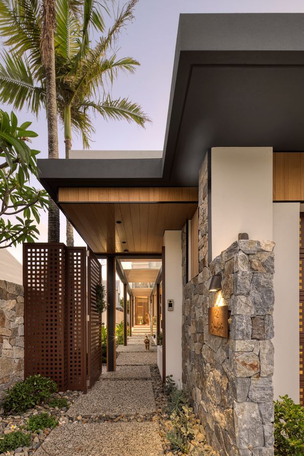 A narrow landscaped entry passage leading toward a modern residence, framed by dark timber and concrete roof overhangs, warm timber soffits, textured natural-stone walls, and perforated brown privacy screens. Palm trees and tropical planting border the gravel walkway, while warm wall lighting and a glimpse of the timber-lined entrance create an inviting resort-like atmosphere.