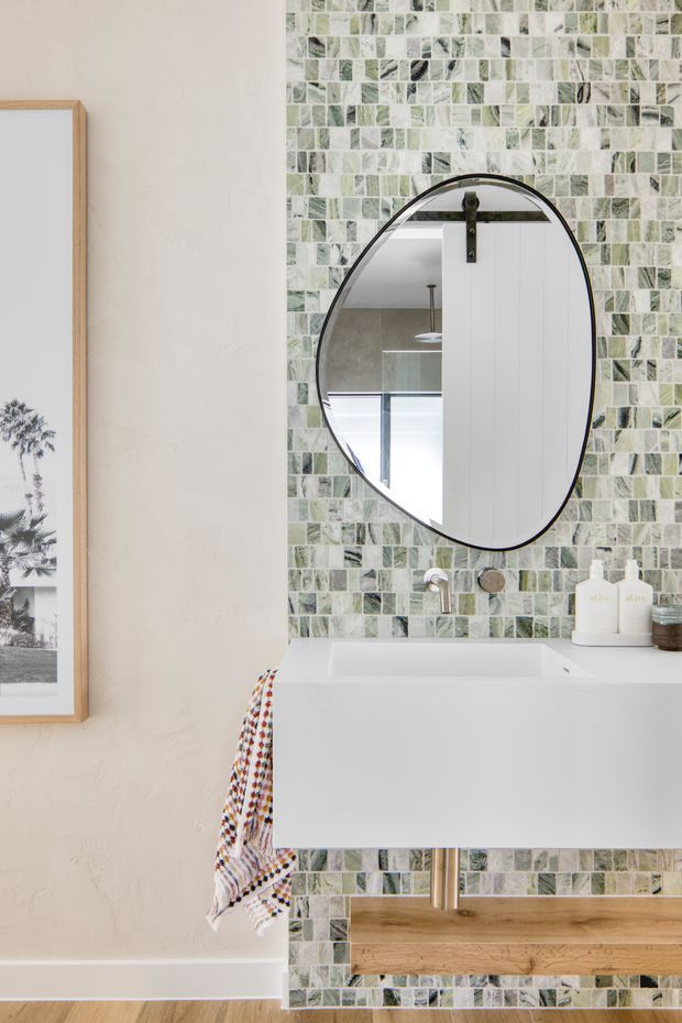 A bright, contemporary bathroom vanity featuring a wall-mounted white basin cabinet, a timber open shelf below, a rounded black-framed mirror, and a green, white, and grey mosaic tile feature wall. The adjacent wall is textured off-white, with a framed artwork and a patterned towel adding small decorative accents.