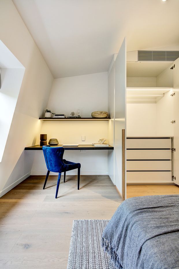 A compact, bright bedroom with a built-in study nook and open wardrobe. The room features pale timber flooring, warm white walls and ceiling, integrated shelving with concealed under-shelf lighting, a dark benchtop desk, and a vivid blue upholstered chair. A bed with grey textured bedding and a woven rug occupies the foreground, while the open wardrobe provides built-in drawers and hanging storage.