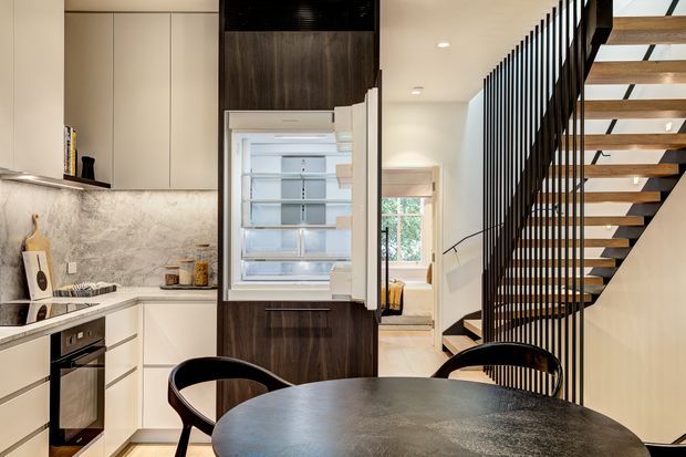 A contemporary open-plan kitchen and dining area beneath a striking open-riser staircase. The kitchen features pale flat-panel cabinetry, a veined stone-look splashback, dark timber cabinetry around the integrated refrigerator, and stainless-steel appliances. A round dark dining table and curved black chairs occupy the foreground, while a bright hallway and window are visible beyond the stair.