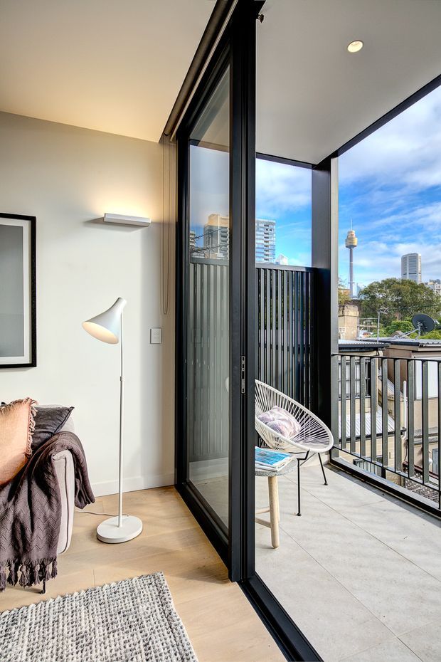A bright contemporary apartment interior opening onto a balcony through full-height black-framed sliding glass doors. The room has pale timber flooring, white walls and ceiling, a compact lounge area, a floor lamp and a textured rug. The balcony features light tiled flooring, black metal balustrades and a woven outdoor chair, with a clear urban skyline visible beyond.